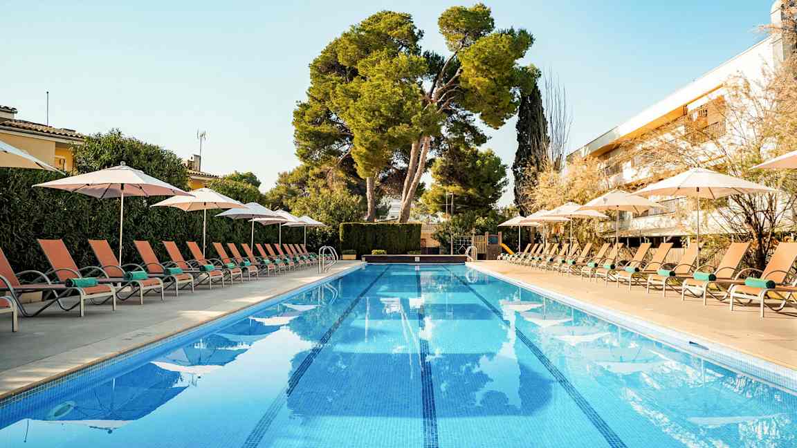 En pool med solstolar och parasoll på båda sidor, omgiven av grönska och med en klar blå himmel. (AI-genererad text)