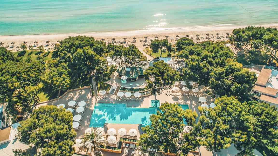 Flygfoto av en strandresort med pool, omgiven av träd och stranden i bakgrunden. (AI-genererad text)