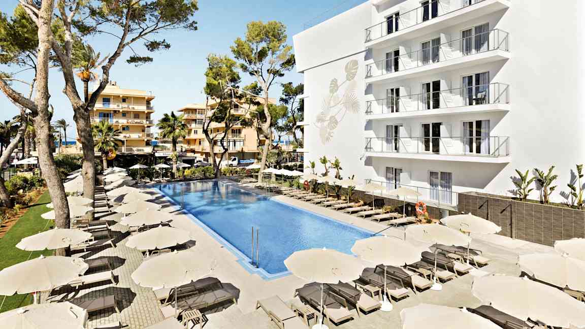 Bild av ett hotellområde med en stor pool omgiven av solstolar och parasoller. En vit byggnad med flera balkonger samt träd i bakgrunden. (AI-genererad text)