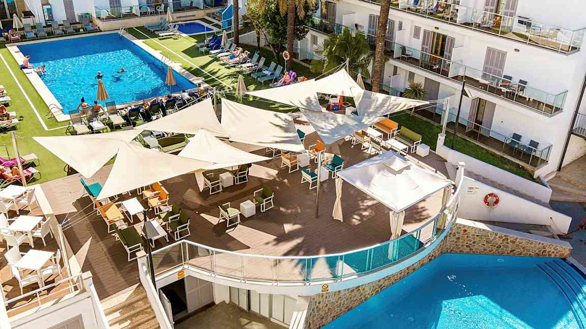 En bild av en hotellresort med en större pool omgiven av solstolar och parasoller. I förgrunden finns en utomhusbar med mängder av sittplatser under skuggande segeldukar. (AI-genererad text)