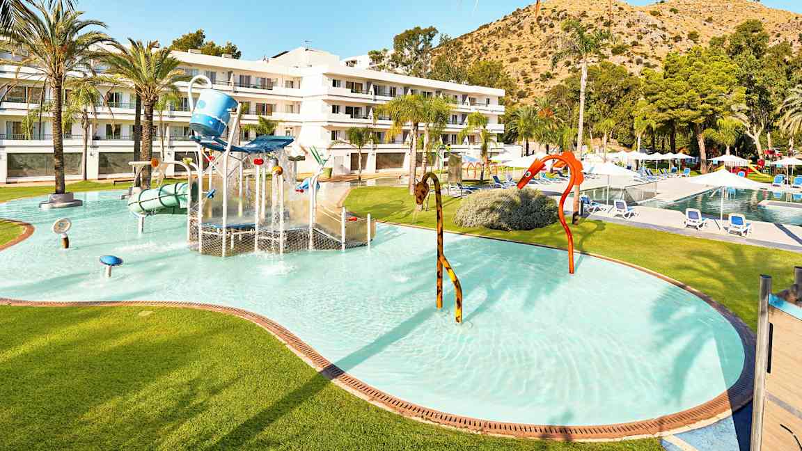 Barnpool med vattenlek och hotellbyggnad i bakgrunden. (AI-genererad text)