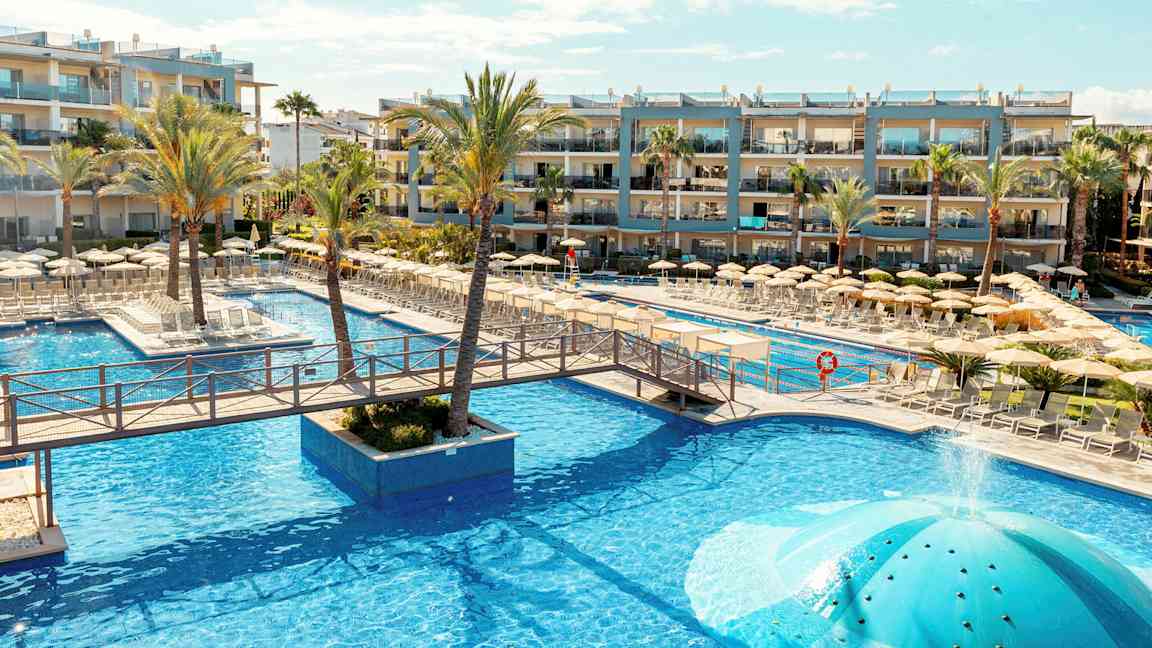 Poolområde vid ett hotell med solstolar och parasoller, omgivet av palmer och byggnader. (AI-genererad text)