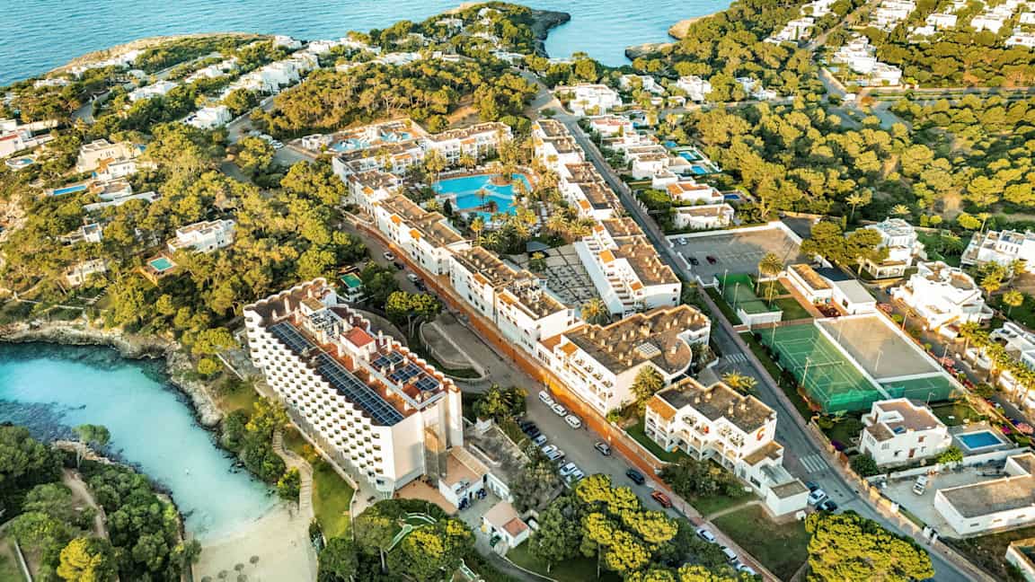 Flygfoto av en semesteranläggning vid kusten med hotell, pooler och gröna områden. (AI-genererad text)