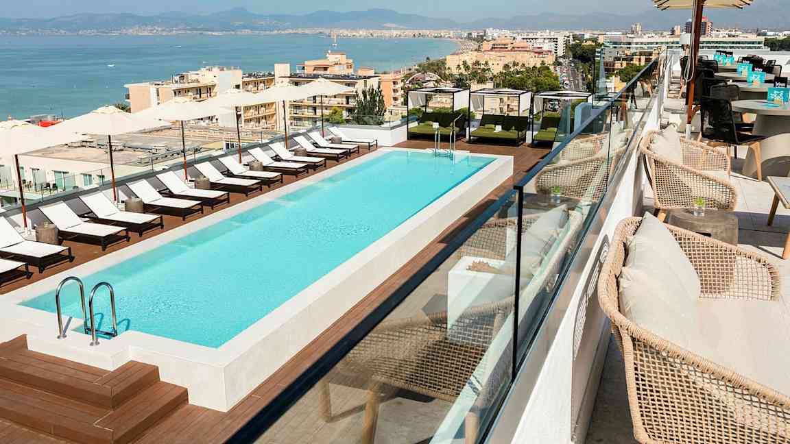 Takvåning med en utomhuspool omgiven av solstolar och skuggsegel, med utsikt över en kuststad. (AI-genererad text)