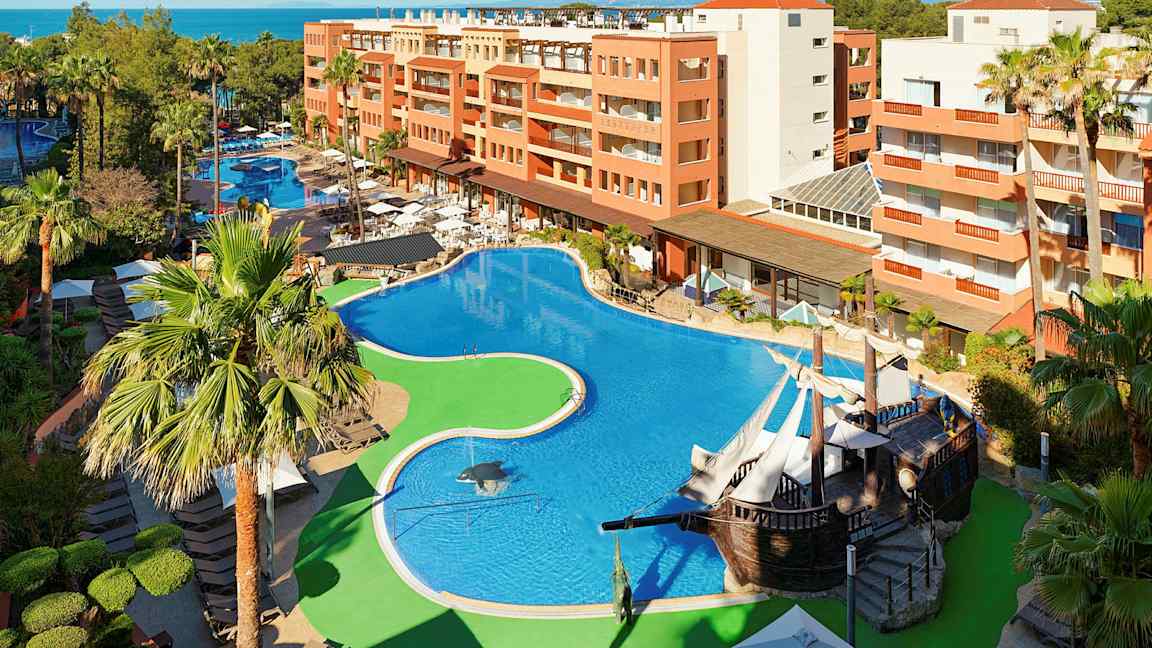 Hotellområde med stor pool, omgiven av palmer och byggnader. (AI-genererad text)