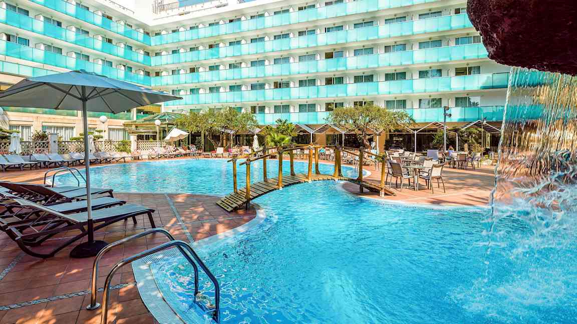 En utomhuspool vid en hotellbyggnad med solstolar, bord och parasoller. En liten bro över poolen och en del av en vattenfallstruktur syns. (AI-genererad text)