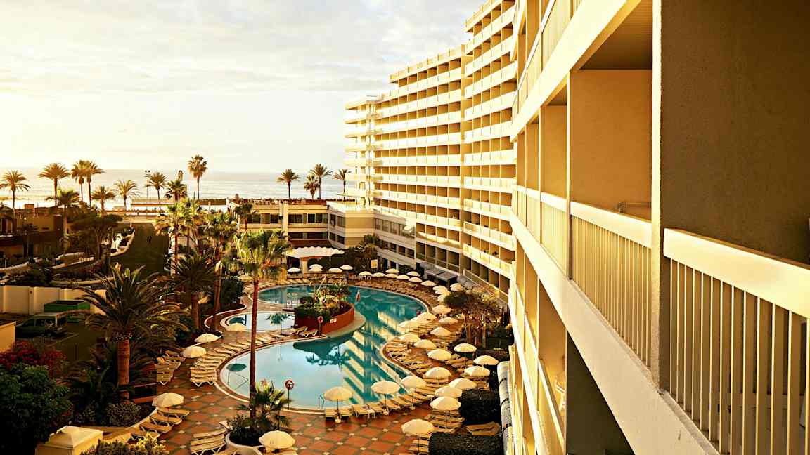 En vy över en hotellpool omgiven av solstolar och parasoller, med palmträd vid sidan och havet i bakgrunden. (AI-genererad text)