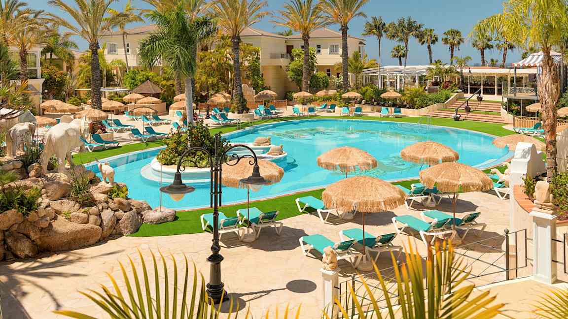 En bild av en lyxig semesteranläggning med en pool, omgiven av solstolar och parasoll, omgivna av palmer och byggnader. (AI-genererad text)