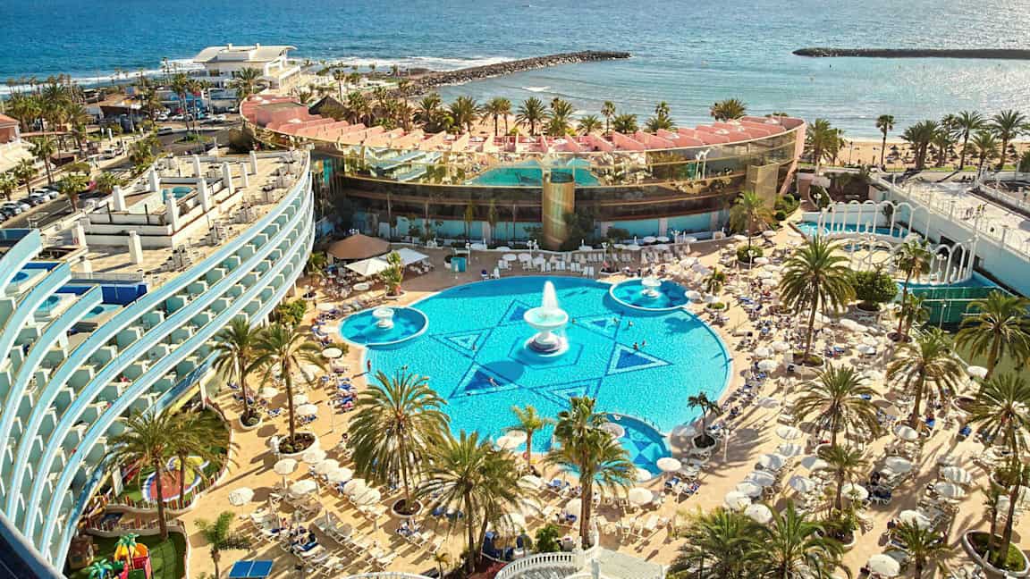 En vy över en resort med poolområde, solstolar och palmer nära havet. (AI-genererad text)