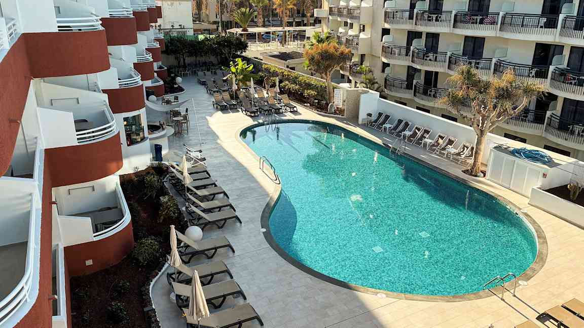 Ett hotell med flera balkonger och en formad pool omgiven av solstolar och parasoller. (AI-genererad text)