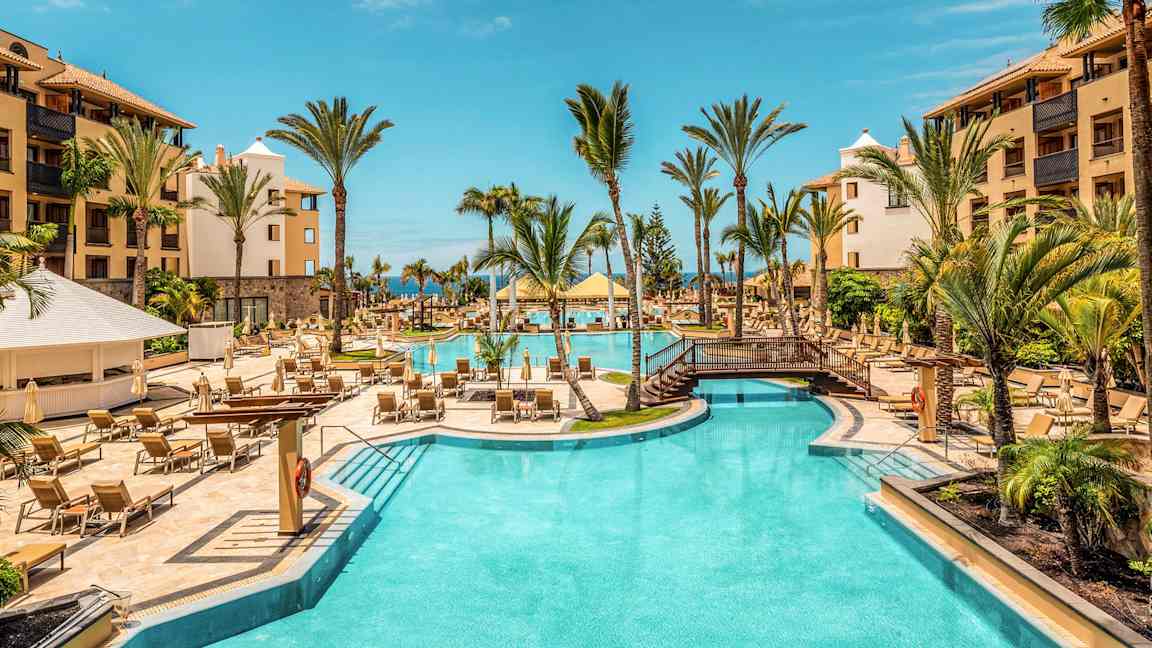 Hotellpool utomhus med palmer och solstolar, omgiven av byggnader. (AI-genererad text)