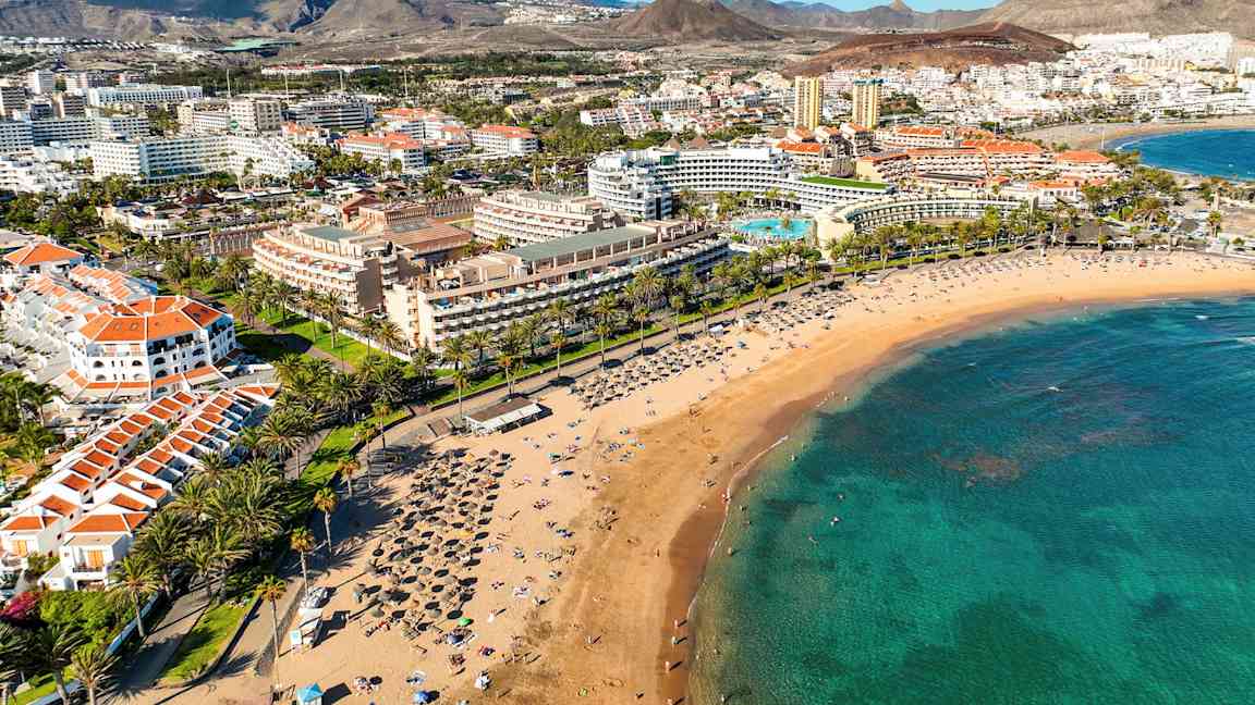 En sandstrand med solstolar och parasoller, omgiven av palmer och hotellbyggnader på ett turistområde. (AI-genererad text)