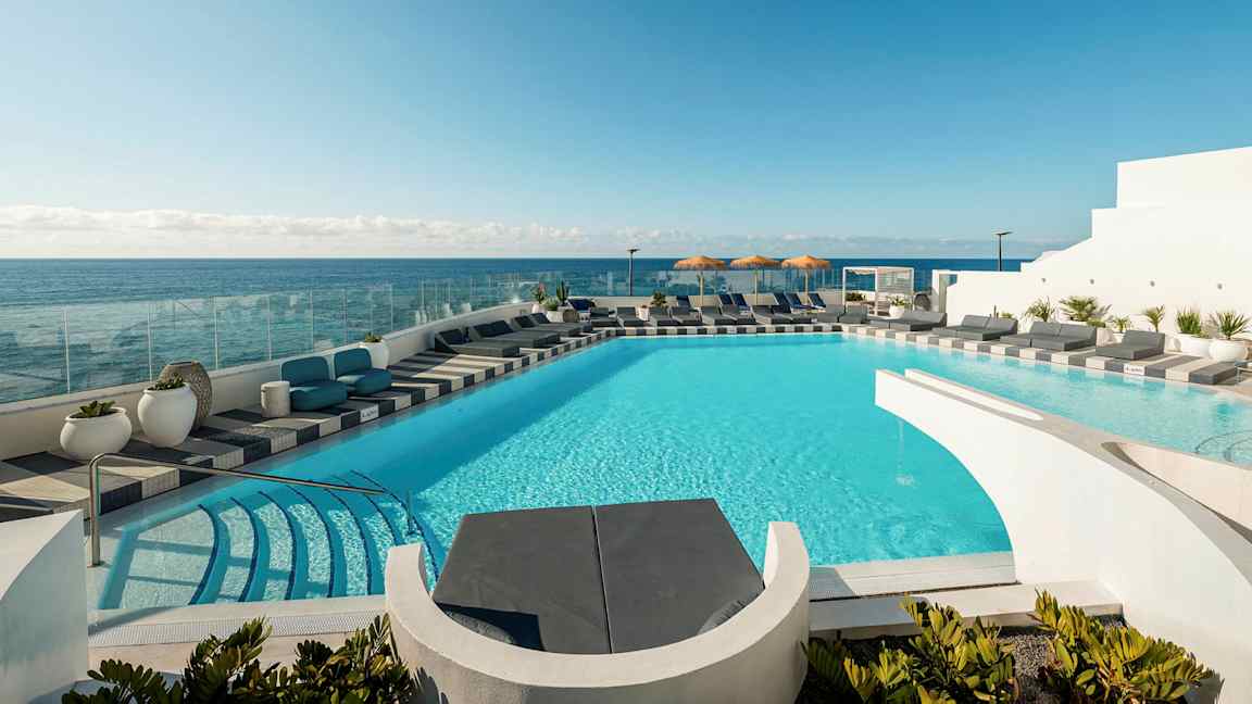 En pool med turkost vatten omgivet av solstolar och utsikt över havet. I bakgrunden syns en klar blå himmel. (AI-genererad text)