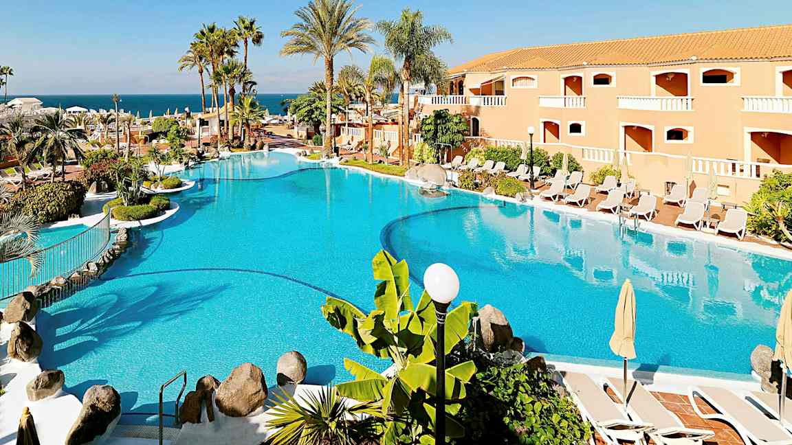 En stor pool omgiven av parasoller, solstolar, palmer och en byggnad i bakgrunden. (AI-genererad text)