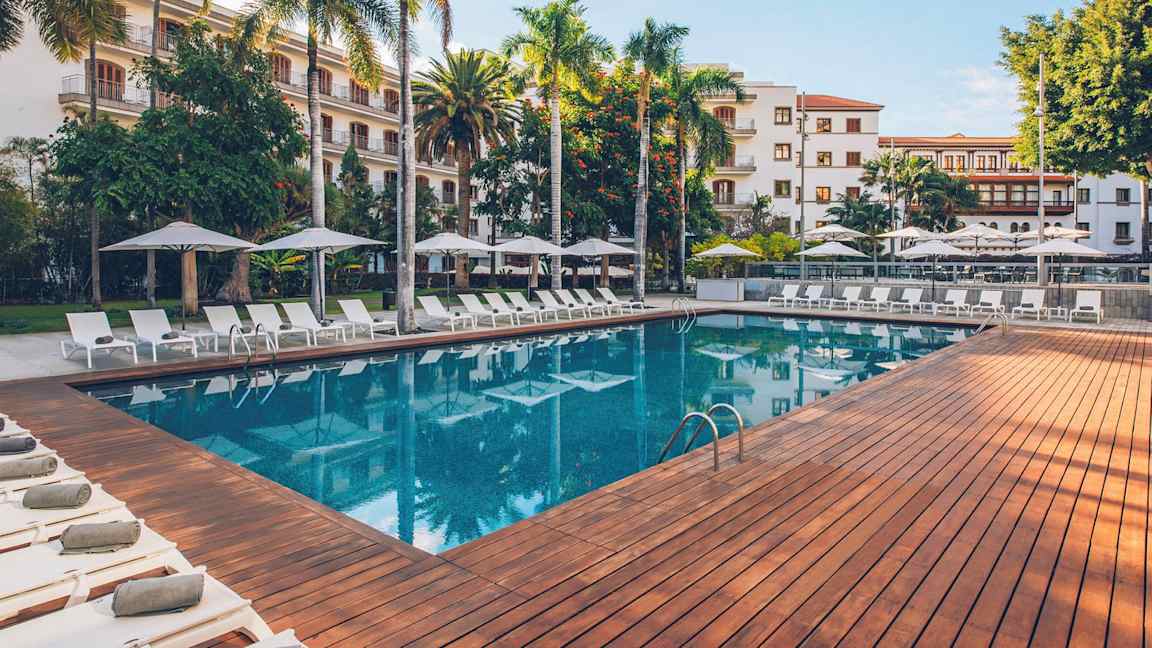 En utomhuspool omgiven av solstolar, parasoll och grönskande träd, med en byggnad i bakgrunden. (AI-genererad text)