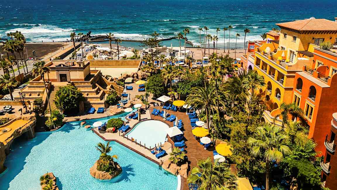 Ett hotellområde med pooler, växtlighet och färgglada byggnader vid havet. (AI-genererad text)
