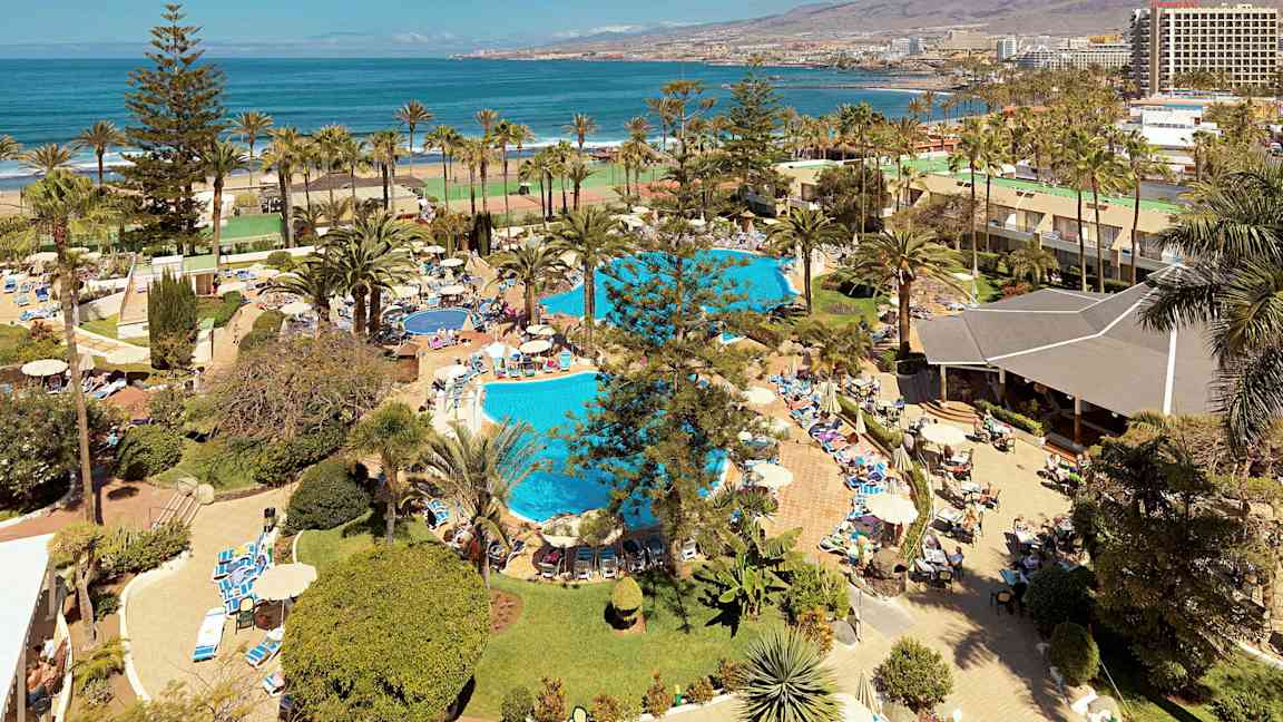 Utsikt över ett semesteranläggning med pooler, palmer och människor som solar och badar nära havet. (AI-genererad text)