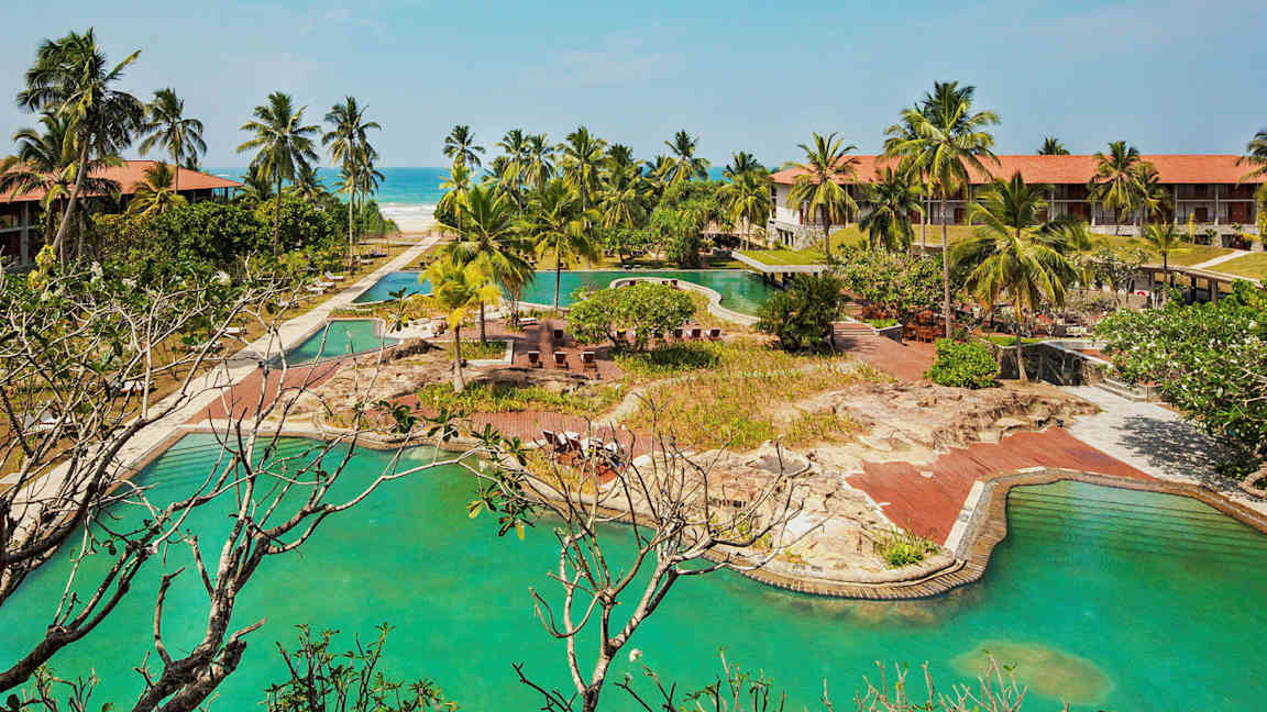 Hotellanläggning med pooler, palmer och en sandstrand i bakgrunden. (AI-genererad text)