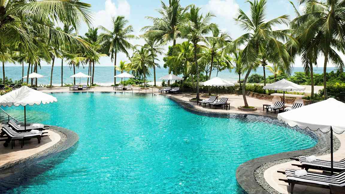 Bild av en pool omgiven av palmer och solsängar med parasoll vid havet. (AI-genererad text)