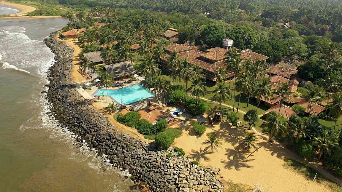 Vy över en kustlinje med ett hotellområde som inkluderar pool, byggnader med tak av tegel och omgivning med grönska och kokospalmer. (AI-genererad text)