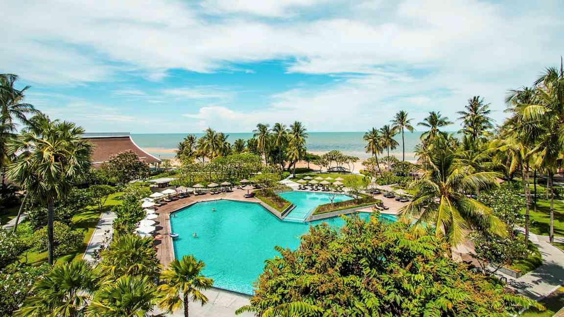 En utomhuspool omgiven av gröna träd och parasoller med utsikt mot havet under en klar blå himmel. (AI-genererad text)