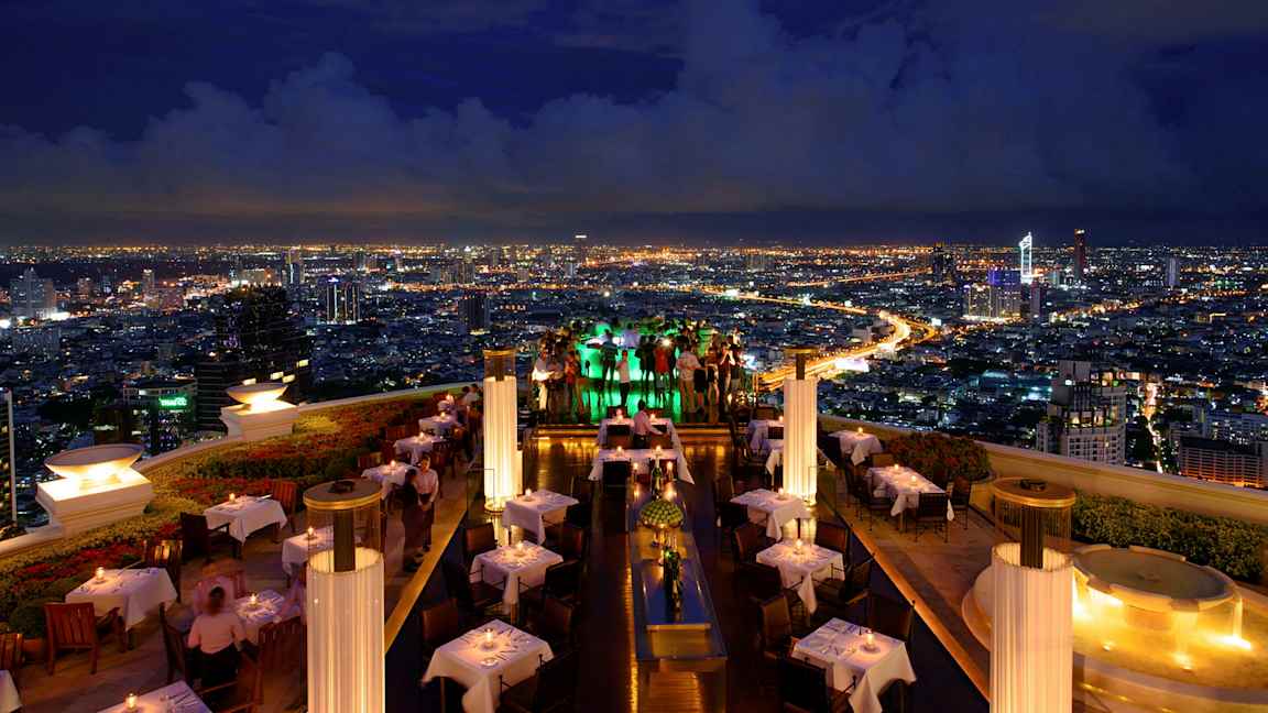 En takterrassrestaurang på kvällen med upplysta bord och utsikt över en stad med lysande ljus. (AI-genererad text)