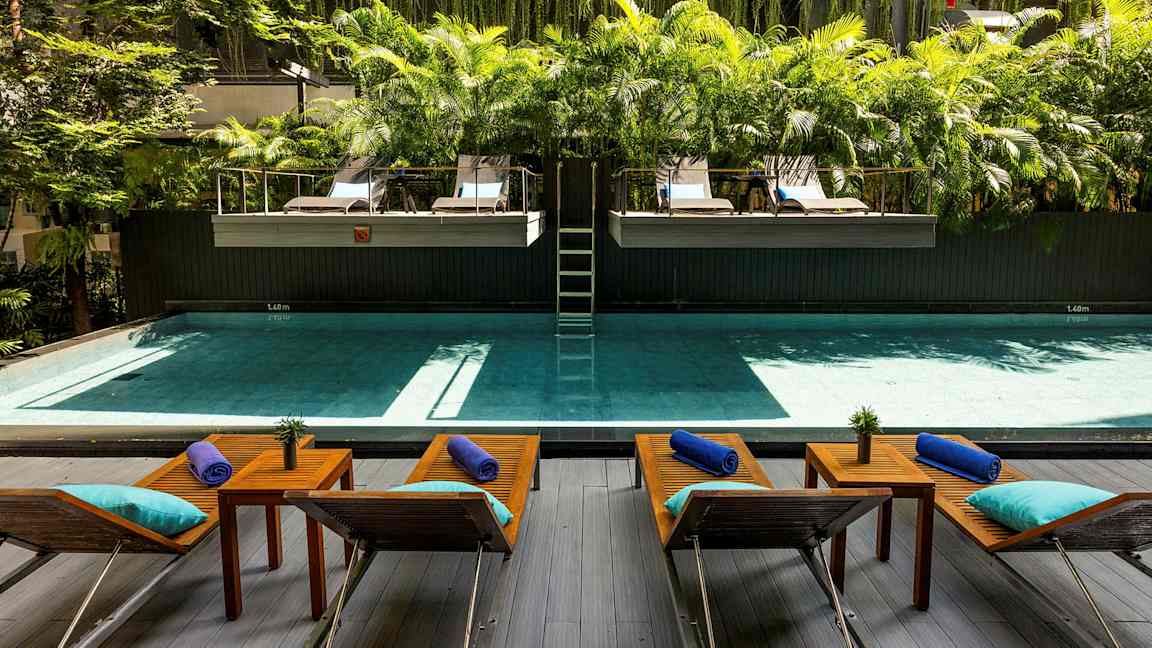 En pool med solstolar och bord framför, omgiven av grönska och palmer. (AI-genererad text)