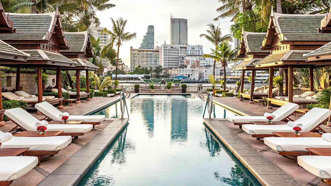 Ett poolområde omgivet av solstolar, palmer och paviljonger i traditionell stil med en stad i bakgrunden. (AI-genererad text)