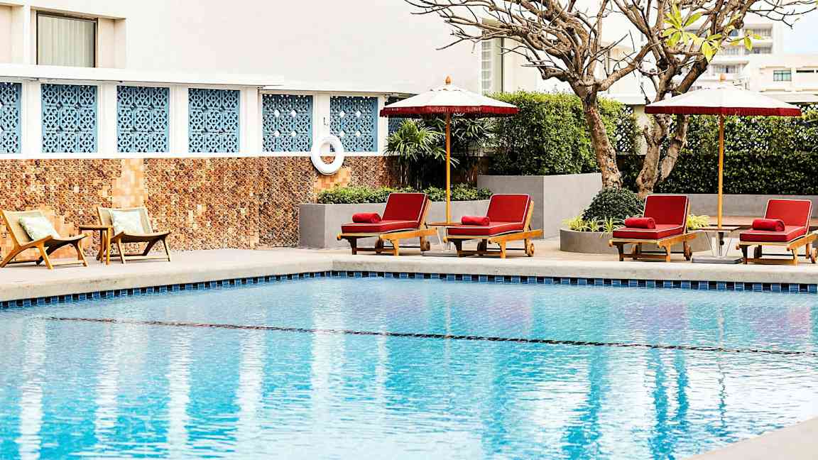 En pool med solstolar och parasoller vid kanten, omgivna av växter och en byggnadsvägg i bakgrunden. (AI-genererad text)