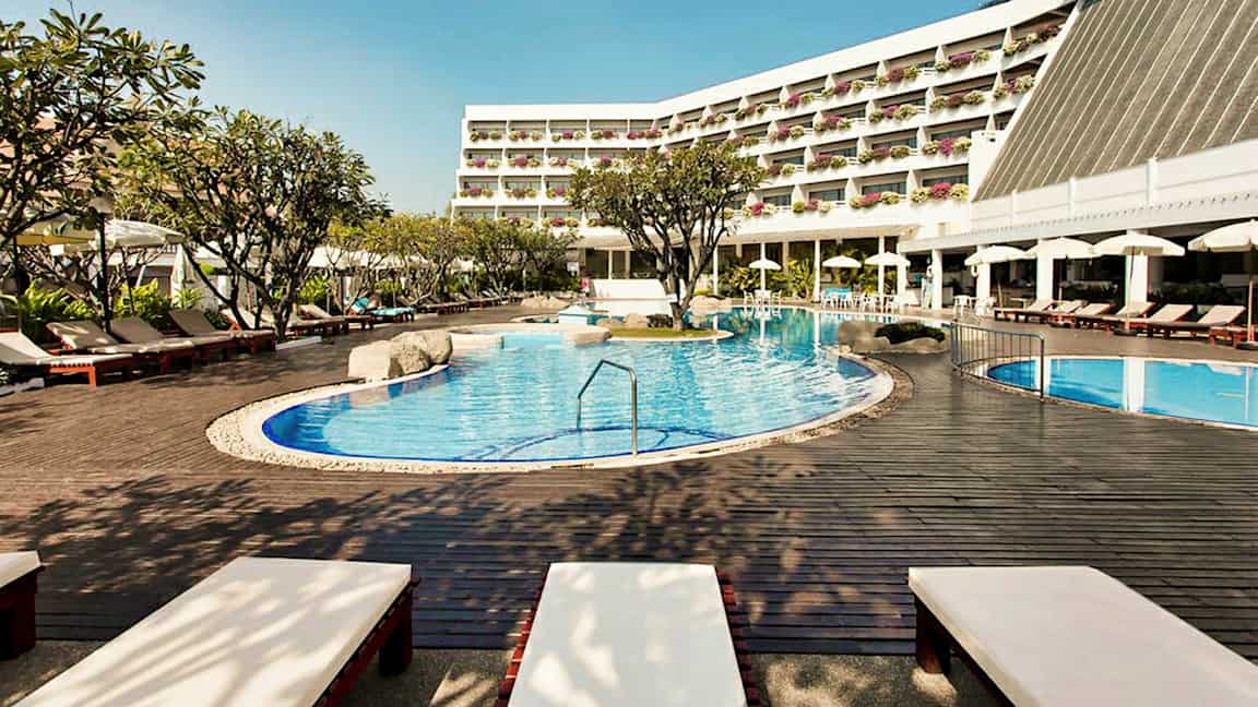 Utomhuspoolområde vid ett hotell med solstolar, parasoller och träd samt en byggnad med balkonger. (AI-genererad text)