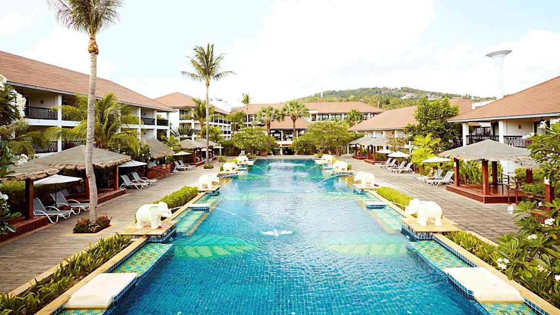 En stor pool på ett semesterortsområde omgiven av palmer, solstolar och bungalowliknande byggnader. (AI-genererad text)