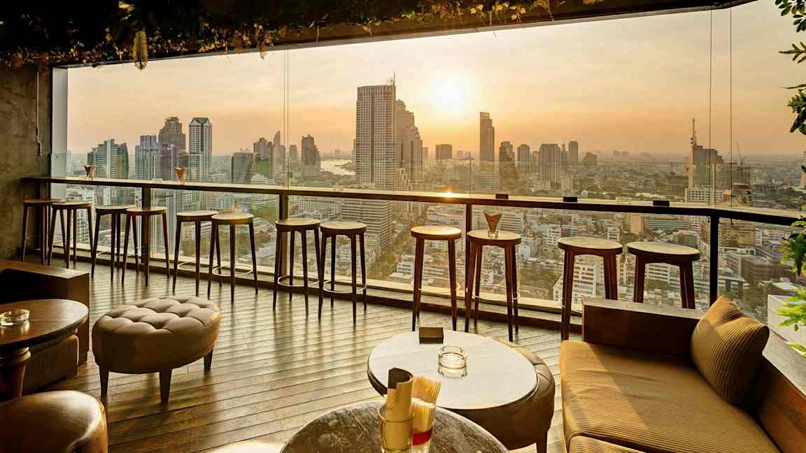 En modern inomhusbar med höga barstolar och bord framför ett stort fönster med utsikt över en stadssilhuett vid solnedgång. (AI-genererad text)