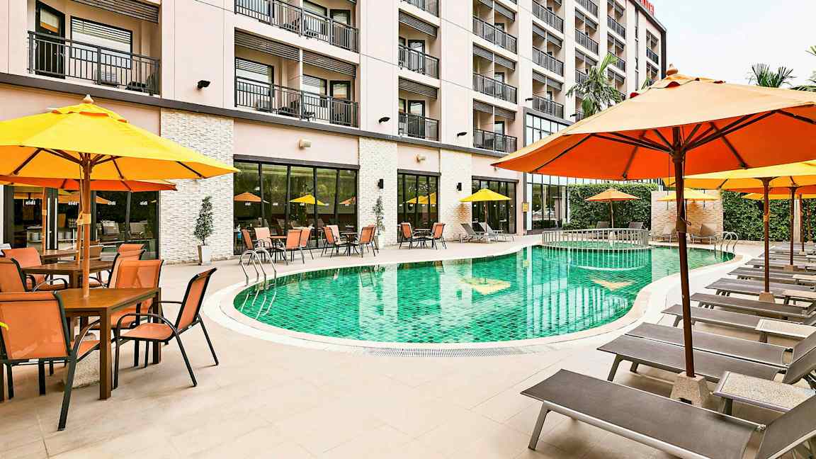 En hotellpool omgiven av solsängar och parasoller, med byggnaden i bakgrunden. (AI-genererad text)