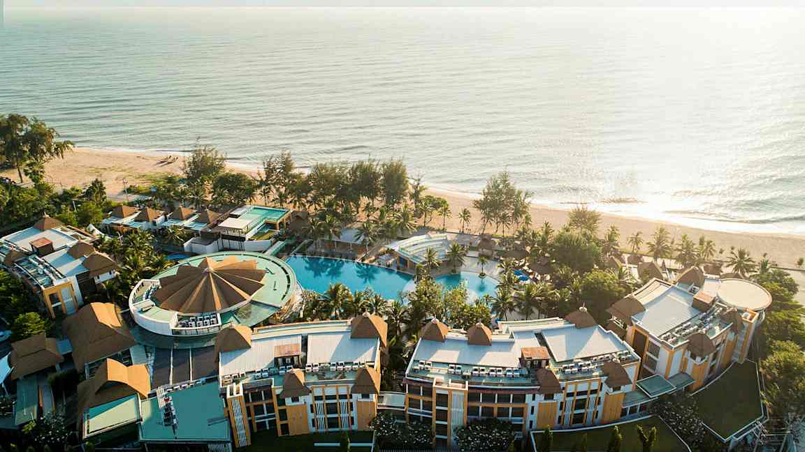 Flygfoto av en resort nära havet med flera byggnader, grönska och en stor pool. (AI-genererad text)