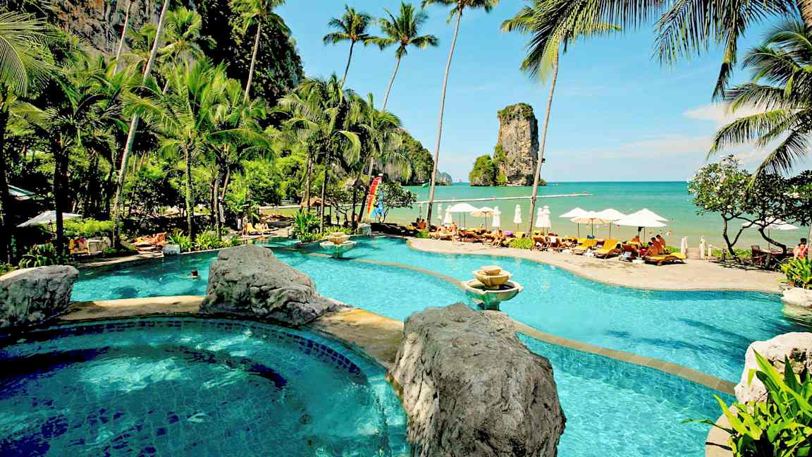 En semesterresort med en pool intill stranden, omgiven av palmer och en storslagen havsutsikt med en klippa vid horisonten. (AI-genererad text)