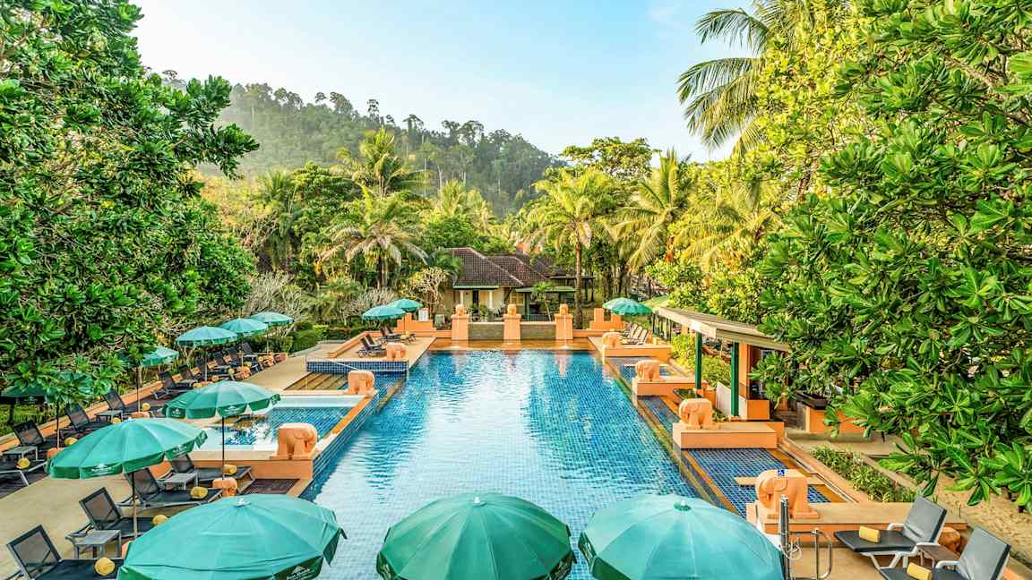 Utomhuspool med blått vatten omgiven av gröna parasoller och solstolar, med grönskande omgivningar och en villa i bakgrunden. (AI-genererad text)