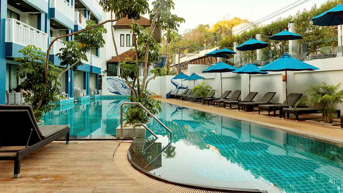 En utomhuspool med solstolar och parasoller, omgiven av vegetation och en hotellbyggnad i flera våningar. (AI-genererad text)