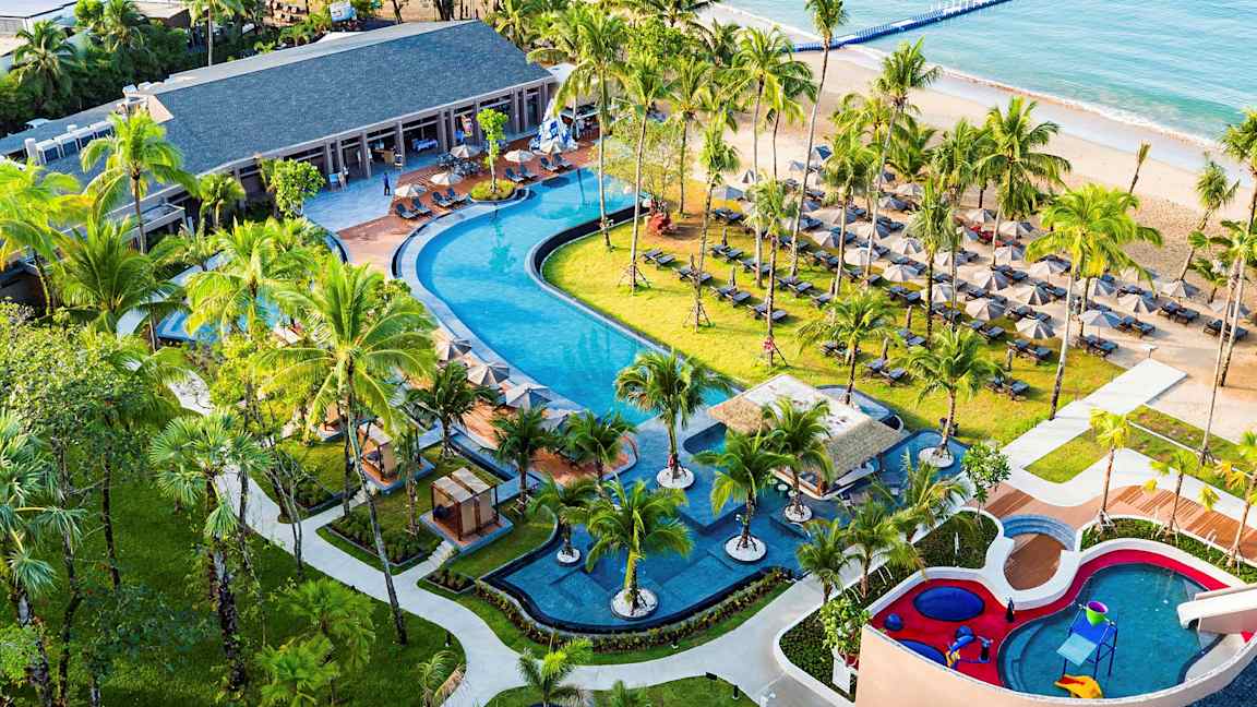 Ett tropiskt semesterområde med en stor pool, omgiven av palmer, solstolar och en barnpool. En sandstrand och havet syns i bakgrunden. (AI-genererad text)