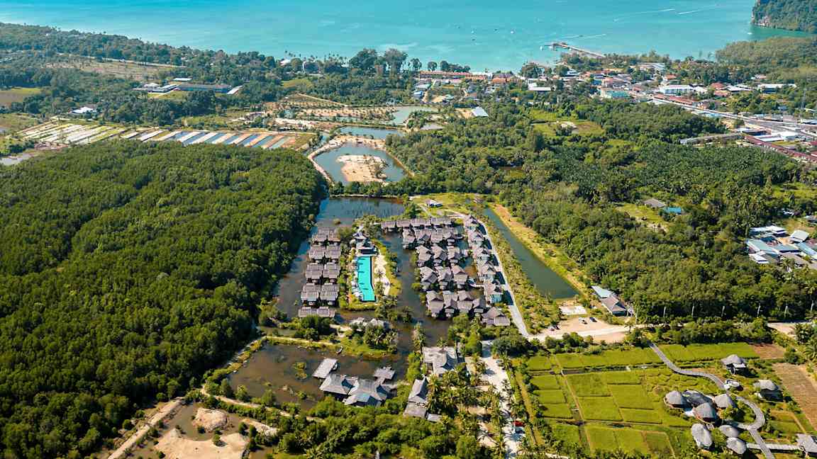 Flygbild över ett resortområde omgiven av vegetation, med en swimmingpool i centrum och havet i bakgrunden. (AI-genererad text)