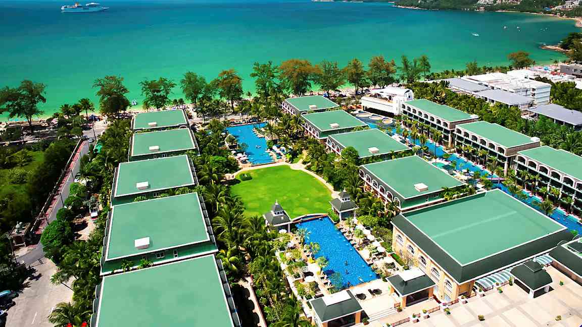 Flygvy över ett semesterhotell med flera byggnader omgivna av grönska och pooler, beläget vid havet. (AI-genererad text)