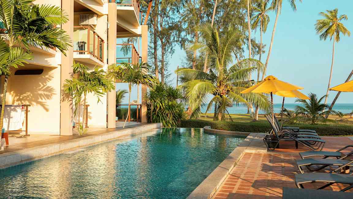 En utomhuspool bredvid ett hotell med balkonger, omgiven av tropiska palmer och gräsyta vid havet. (AI-genererad text)