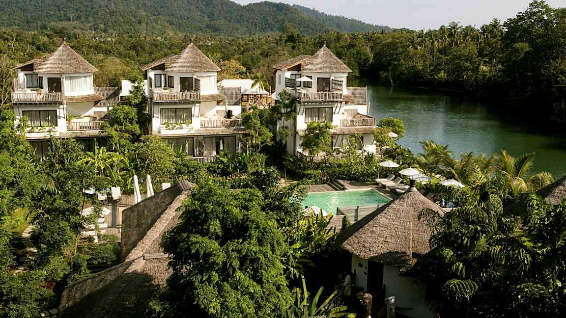 Ett hotellområde med hus i tropisk stil, omgiven av grönska och en flod i bakgrunden. (AI-genererad text)