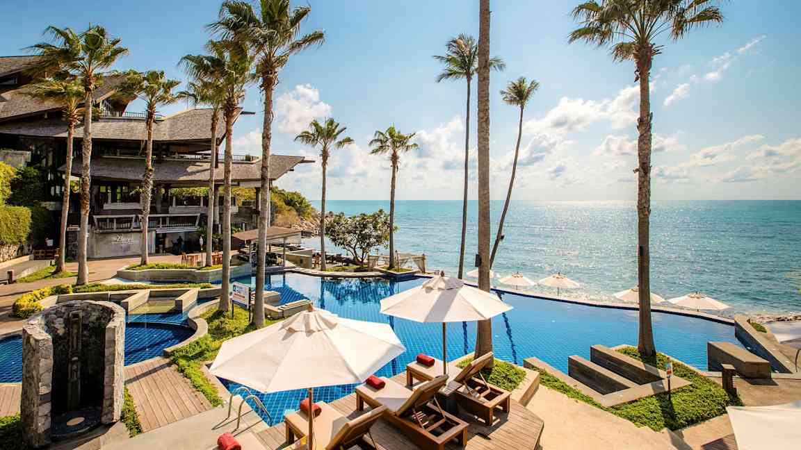 En poolområden vid havet med utsikt över klarblått vatten, omgivna av höga palmer. (AI-genererad text)