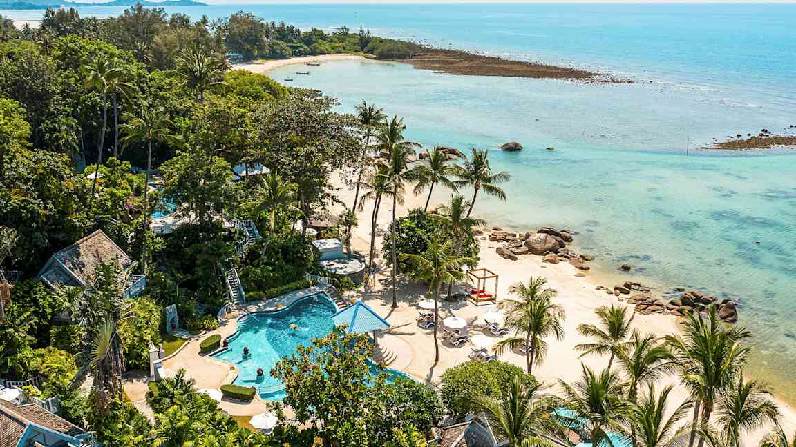 Ett tropiskt landskap med en strand som kantas av palmträd och klarblått vatten. Det finns en pool och gröna områden i förgrunden. (AI-genererad text)