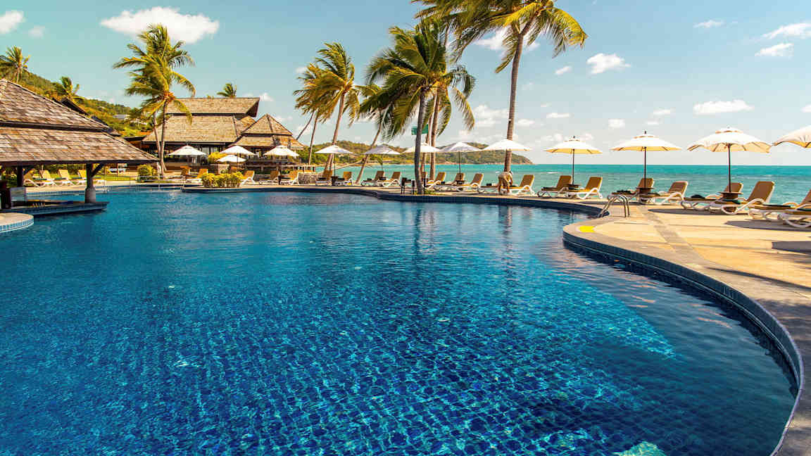 Bild av en utomhuspool omgiven av solstolar och palmträd, med havet i bakgrunden. (AI-genererad text)