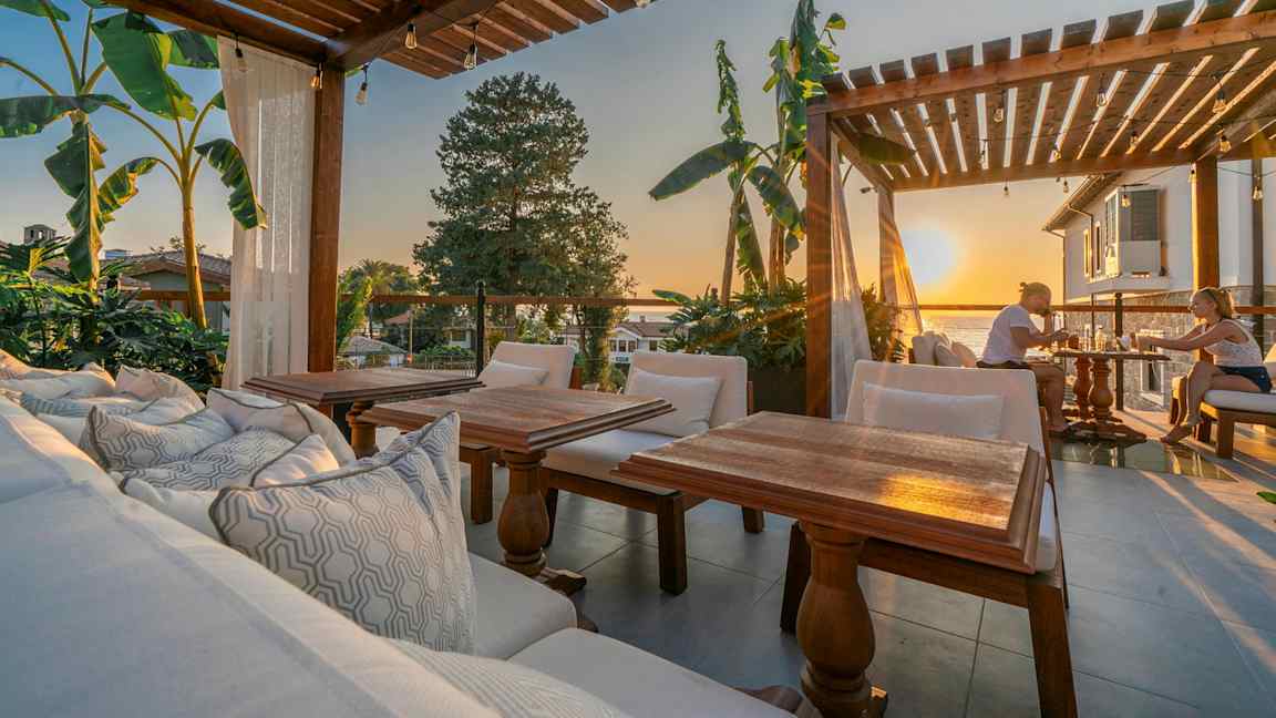 Uteservering med träpergola, sittplatser och utsikt vid solnedgång. (AI-genererad text)