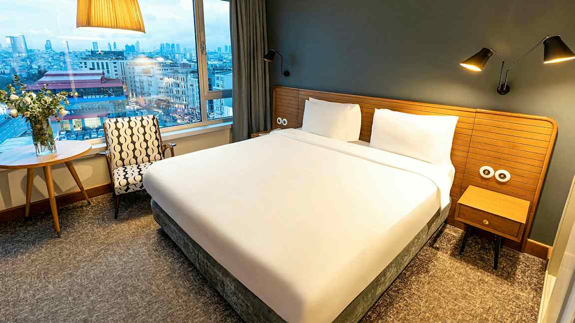 Ett hotellrum med en stor säng, fönster och utsikt över en stad. (AI-genererad text)