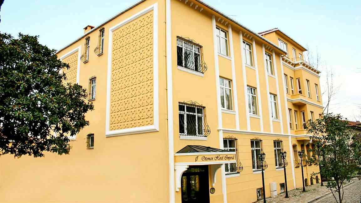 Ett gult hotell med dekorativ fasad, fönster med järngaller och en ingång med takskydd. (AI-genererad text)