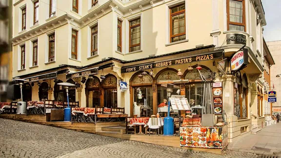 En gata med en byggnad som har restaurang med uteservering, skyltar för mat som kebab och pizza. (AI-genererad text)