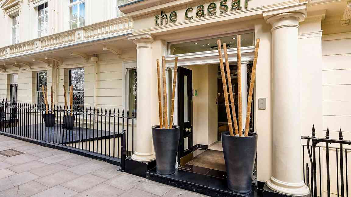 Fasad med ingång till hotell The Caesar, omgiven av svart järnstaket. (AI-genererad text)